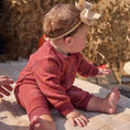 Load image into Gallery viewer, Baby in a red outfit with a headband sitting on a stone surface with hay bales in the background.