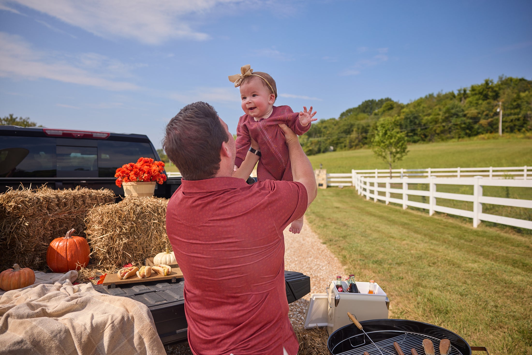 Man lifting a child in a field with hay bales, pumpkins, and a truck in the background.